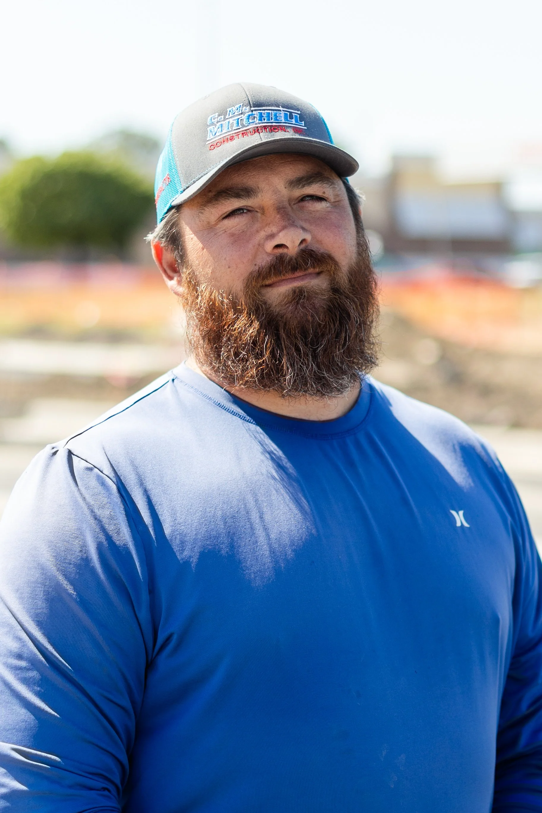 A man with a beard wearing a blue shirt and a gray cap with logos, standing outdoors on a sunny day.