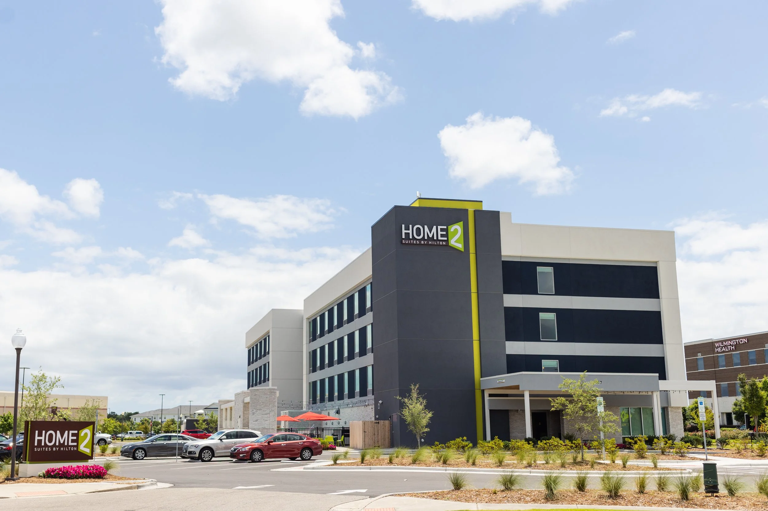 Modern multi-story hotel building labeled 'Home2 Suites by Hilton' with a parking lot in the foreground, cars, landscaped greenery, and clear sky with clouds.