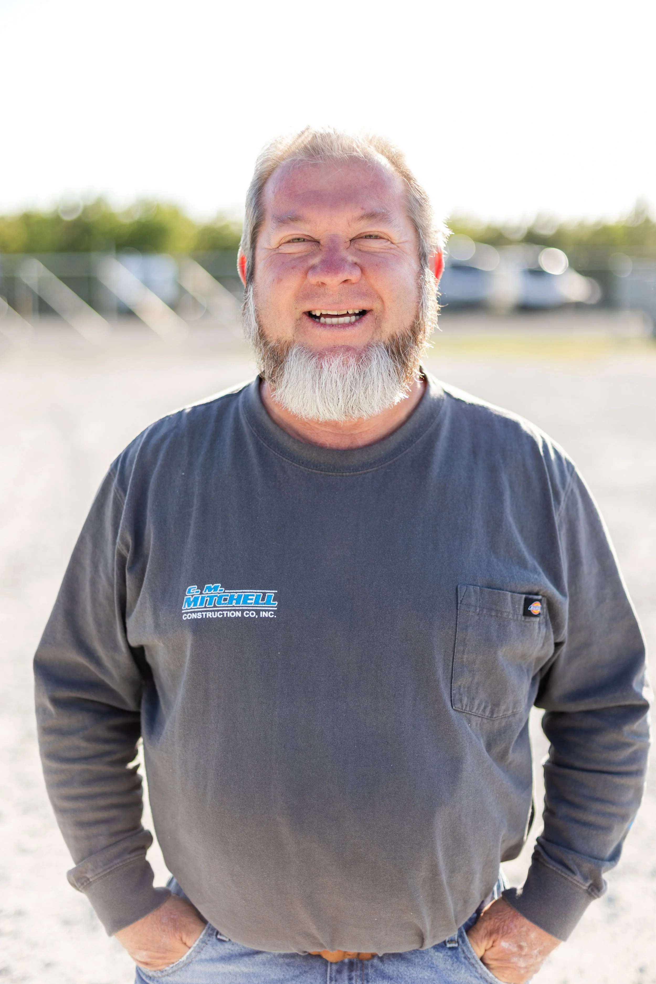 A smiling middle-aged man with a beard wearing a dark gray long-sleeve shirt with a company logo, standing outdoors with blurred background, possibly a parking lot or open area.