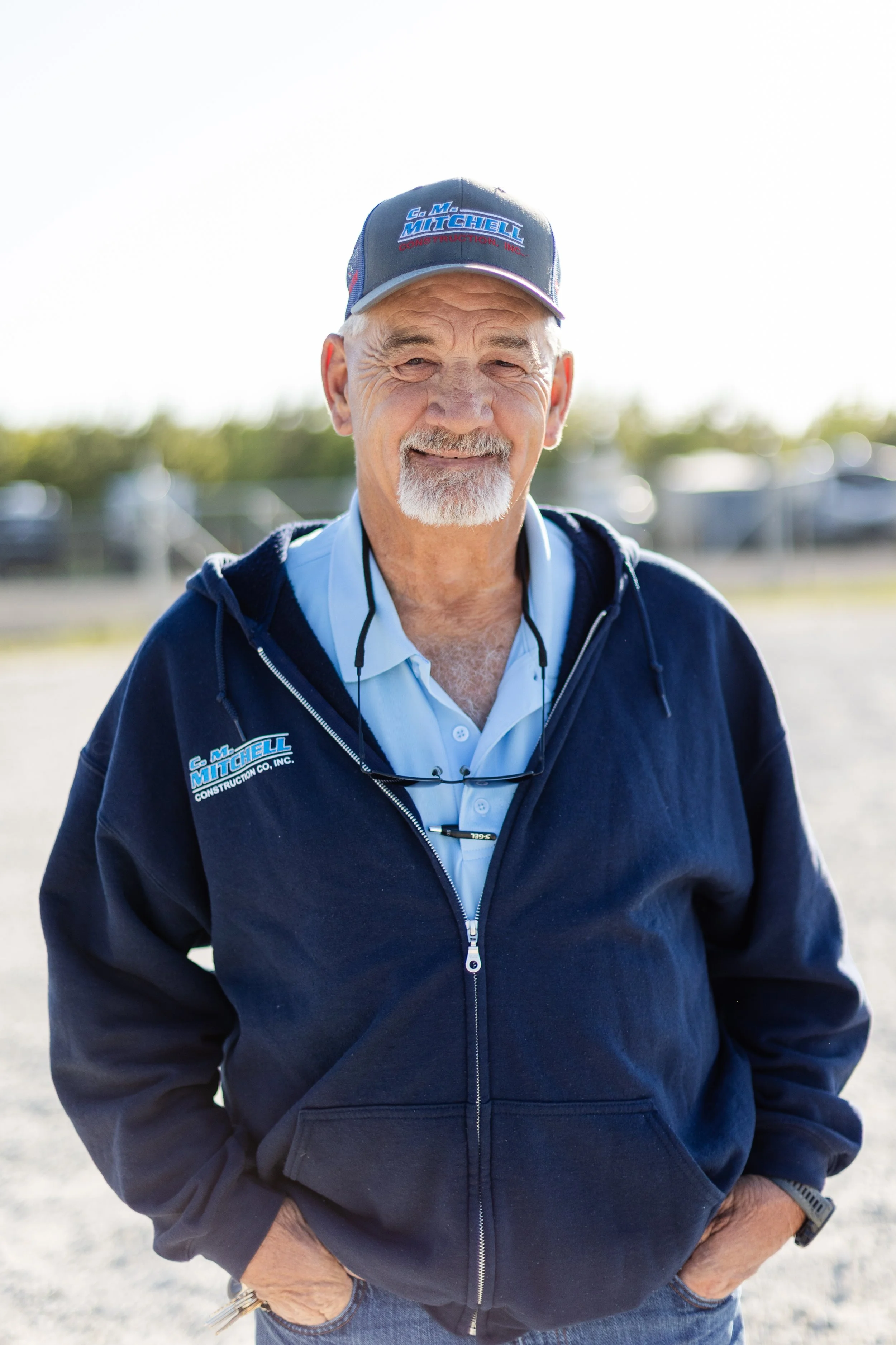 An older man wearing a blue cap and navy jacket with company logos, smiling outdoors on a sunny day with blurred background.