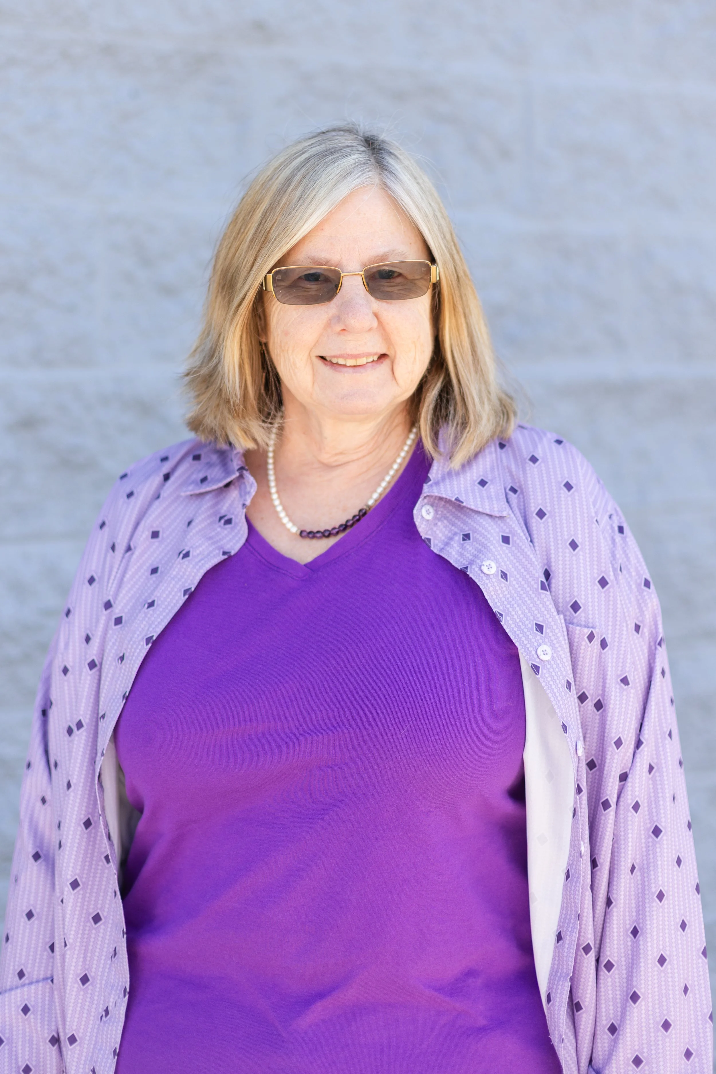 A woman with blonde hair wearing sunglasses, a purple shirt, and a light purple patterned button-up shirt, standing in front of a grey wall.