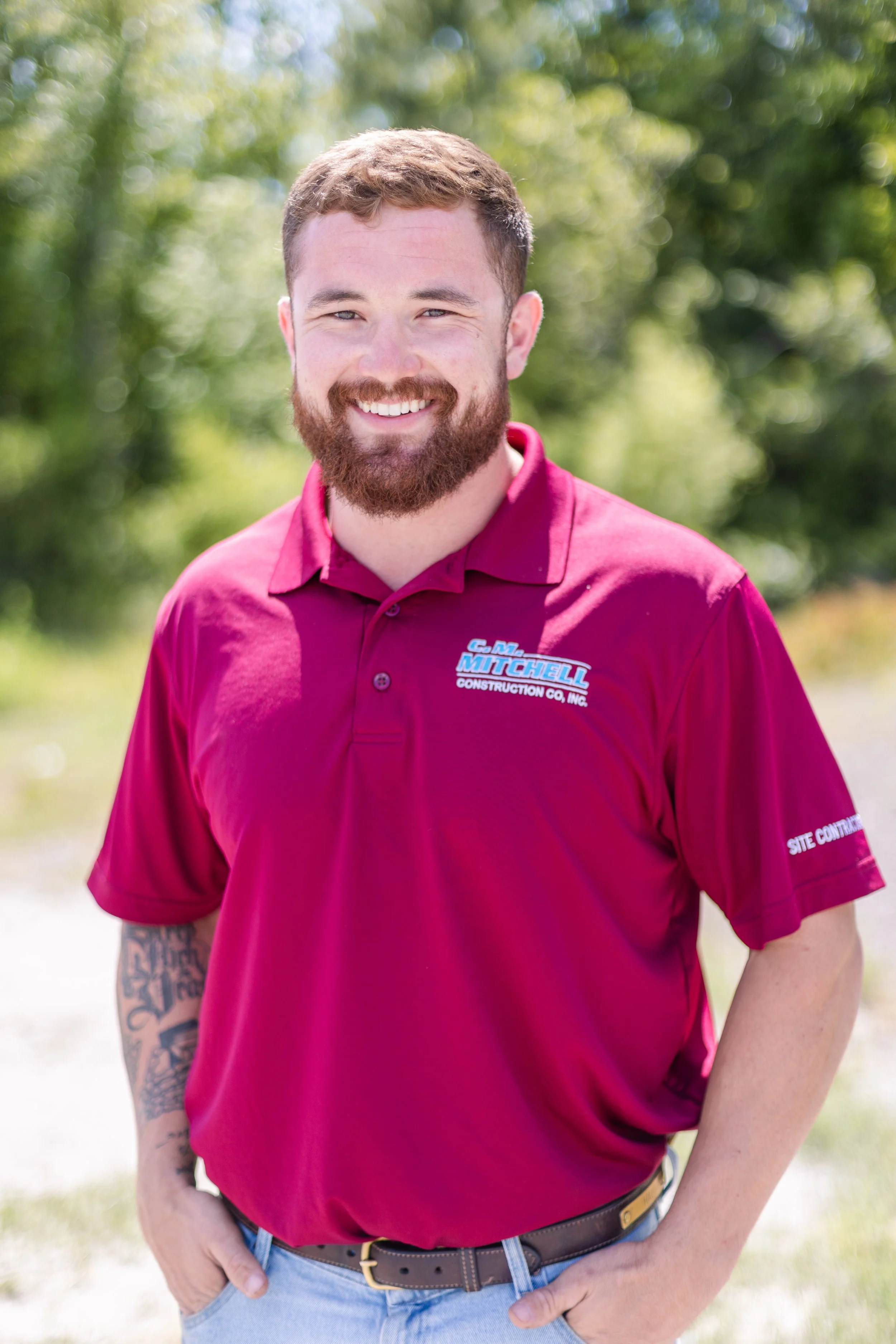 A smiling man with a beard and tattoos wearing a maroon shirt with company logos standing outdoors in a sunny, green environment.