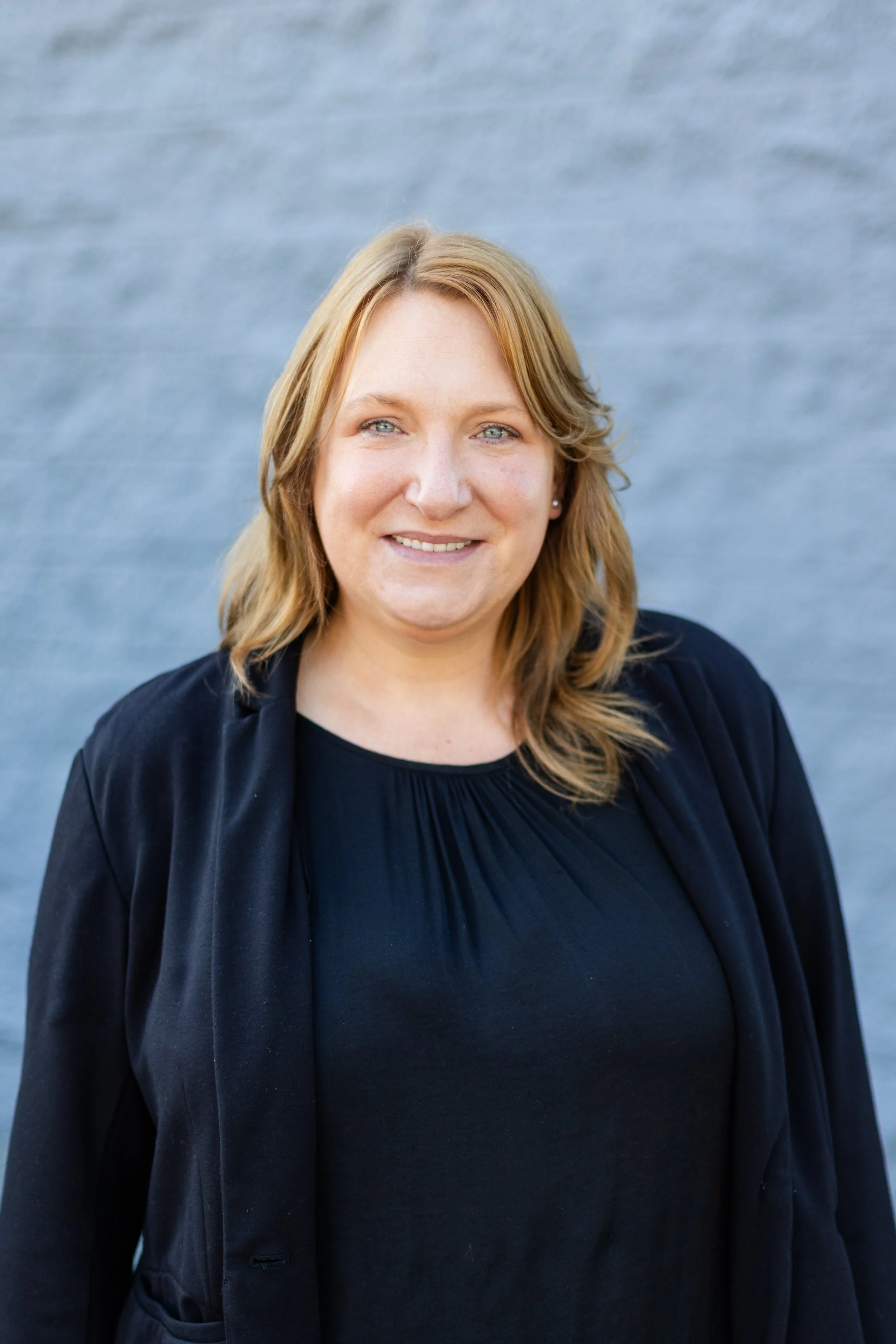 A woman with shoulder-length blonde hair, blue eyes, and fair skin is smiling and wearing a black top and black blazer. The background is a blurred blue body of water.