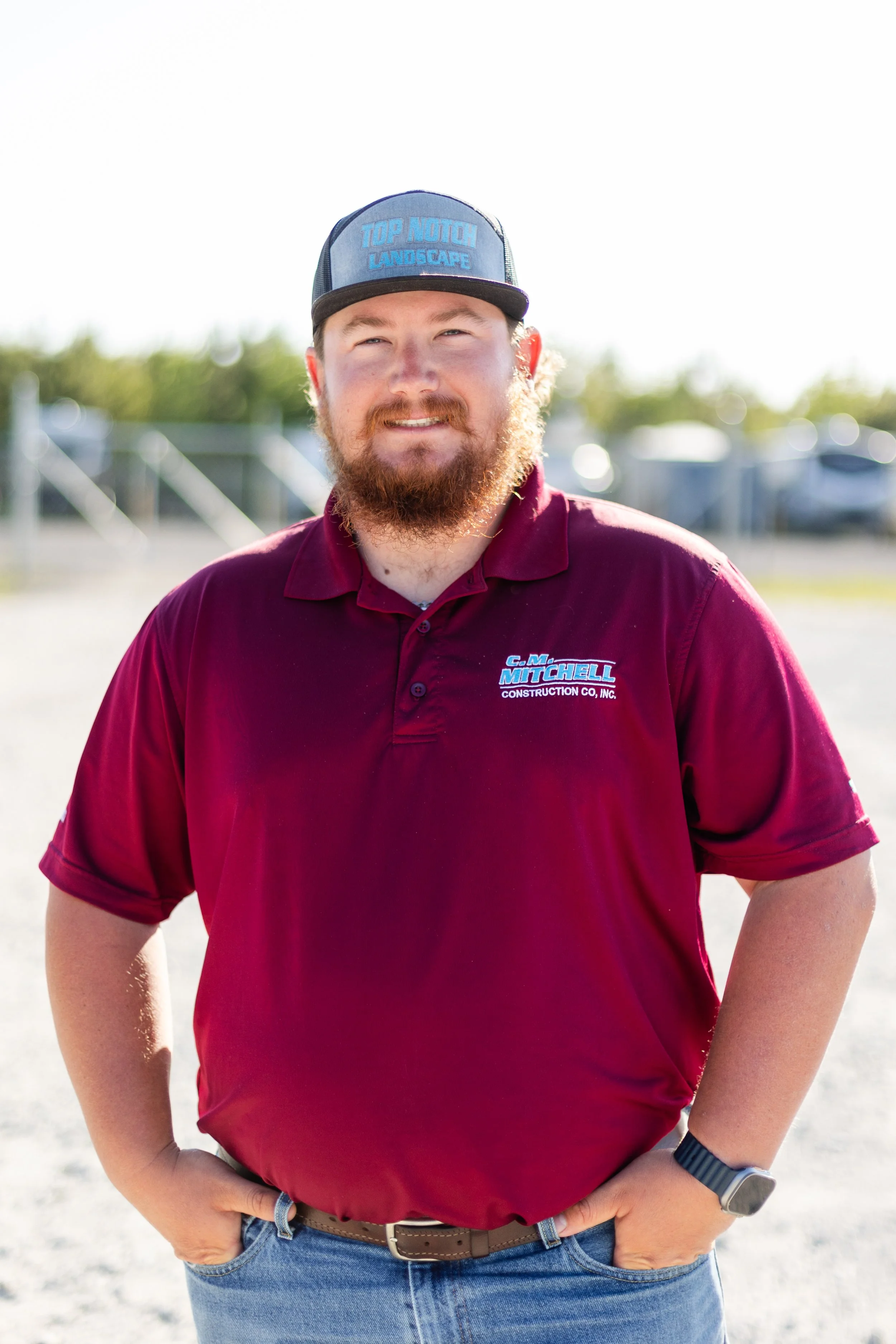Man wearing a maroon polo shirt with logos, a black belt, and a dark baseball cap with blue text, standing outdoors with a bright background. He has a beard and mustache, and he is smiling slightly with one hand on his hip.
