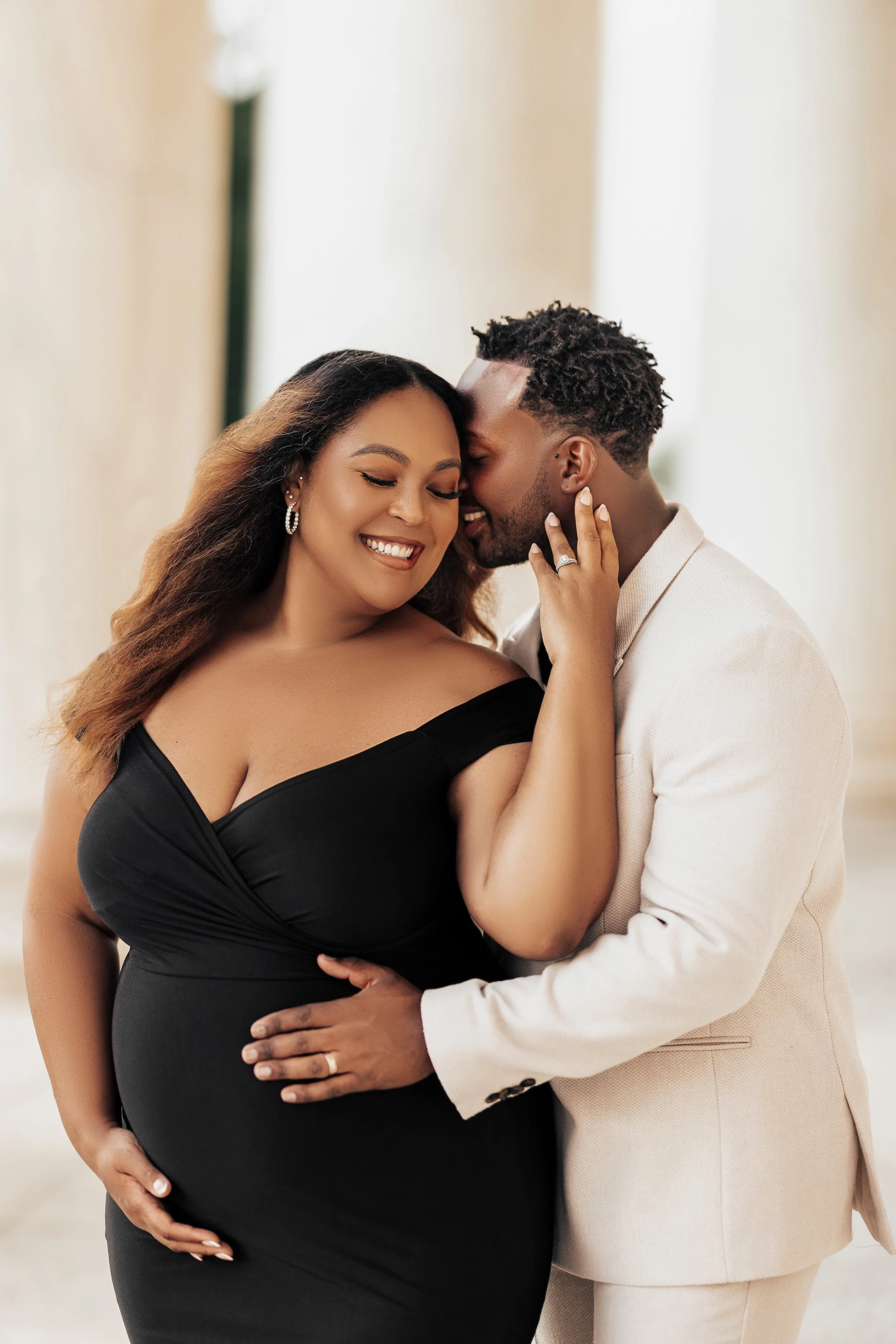 A pregnant woman with long hair in a black dress and earrings smiling with closed eyes, while her partner, a man with short curly hair in a beige suit, kisses her forehead and embraces her, holding her baby bump with one hand and her face with the other, in a softly lit indoor setting.