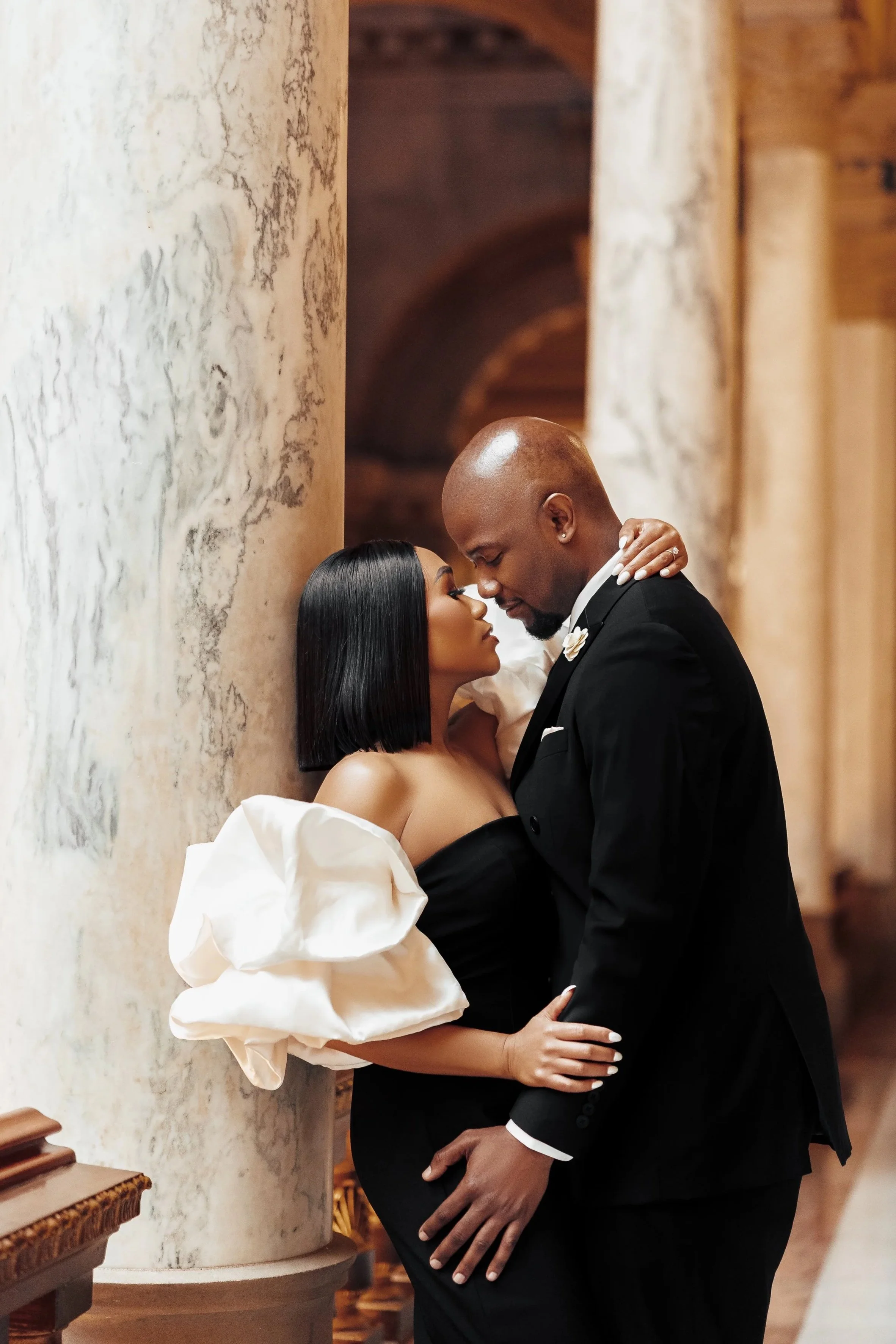 A romantic couple, woman with black hair and man with a shaved head, dressed in formal attire, sharing an intimate moment in an elegant, ornately decorated setting.