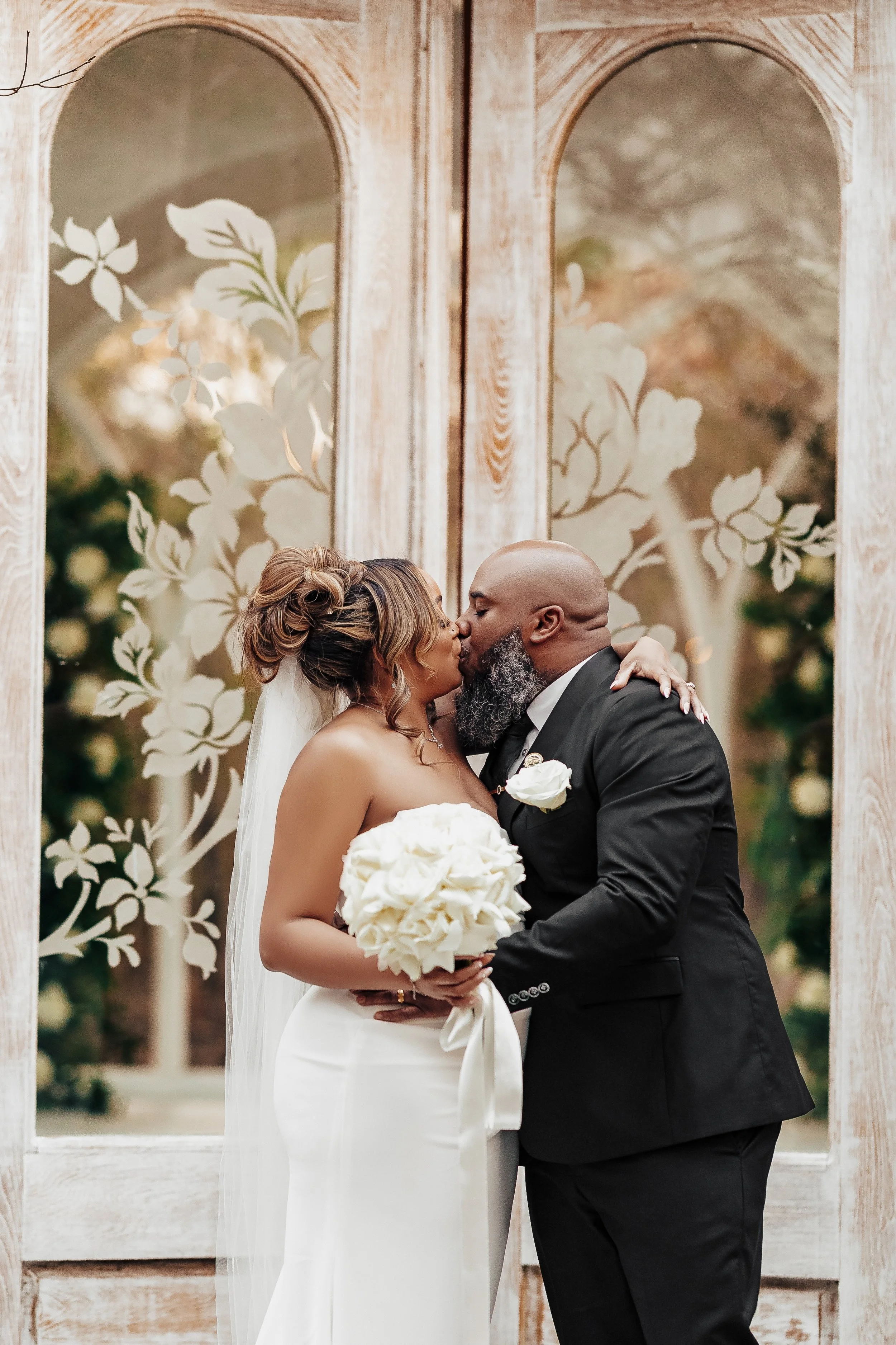 A bride and groom share a kiss during their wedding ceremony in front of decorative wooden arches with floral patterns, outdoors in natural lighting.