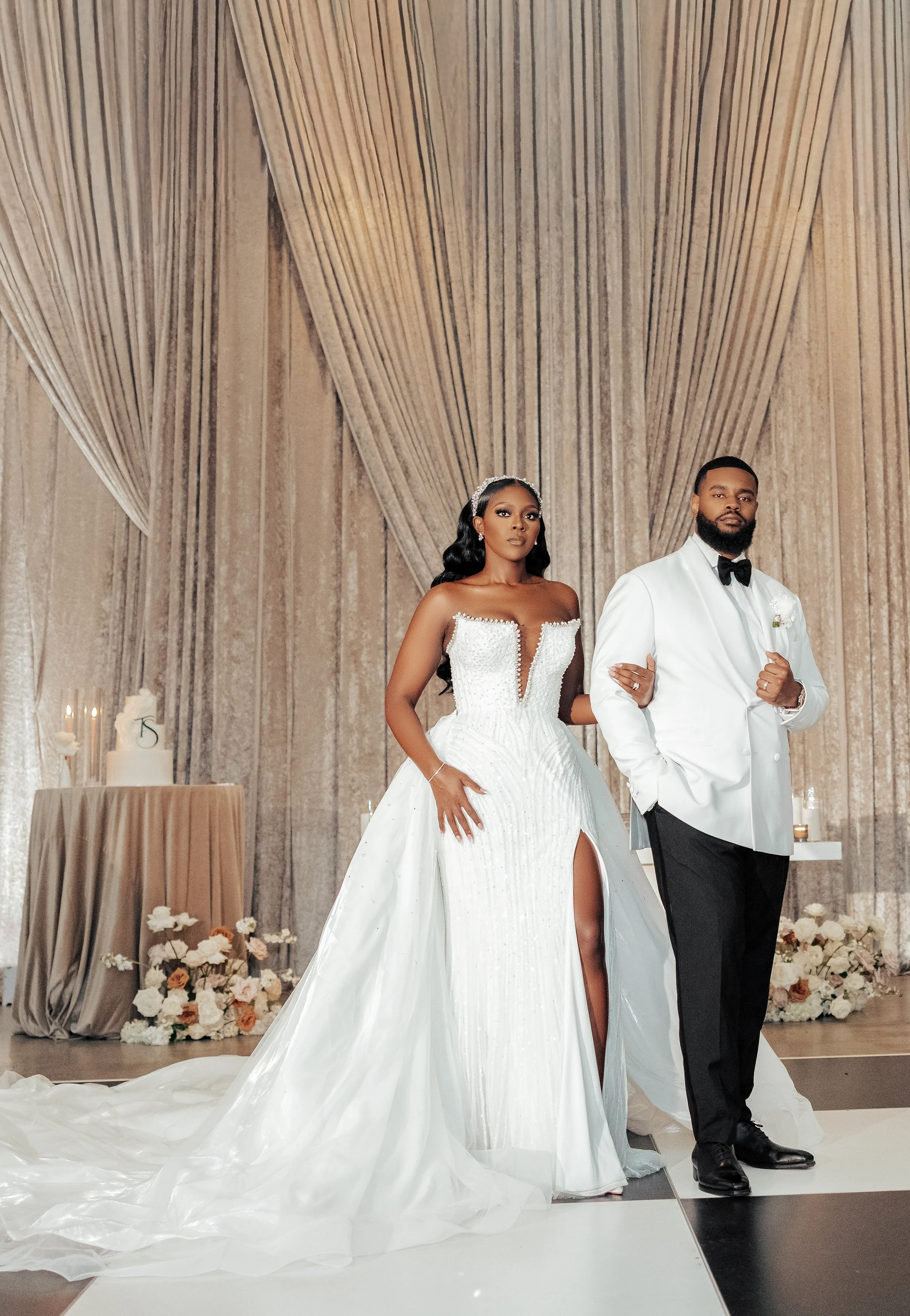 A bride and groom standing side by side in a wedding venue with beige drapes and floral decorations behind them. The bride is in a white wedding gown with a thigh-high slit and a sweetheart neckline, and the groom is in a white tuxedo with a black bow tie and black pants.