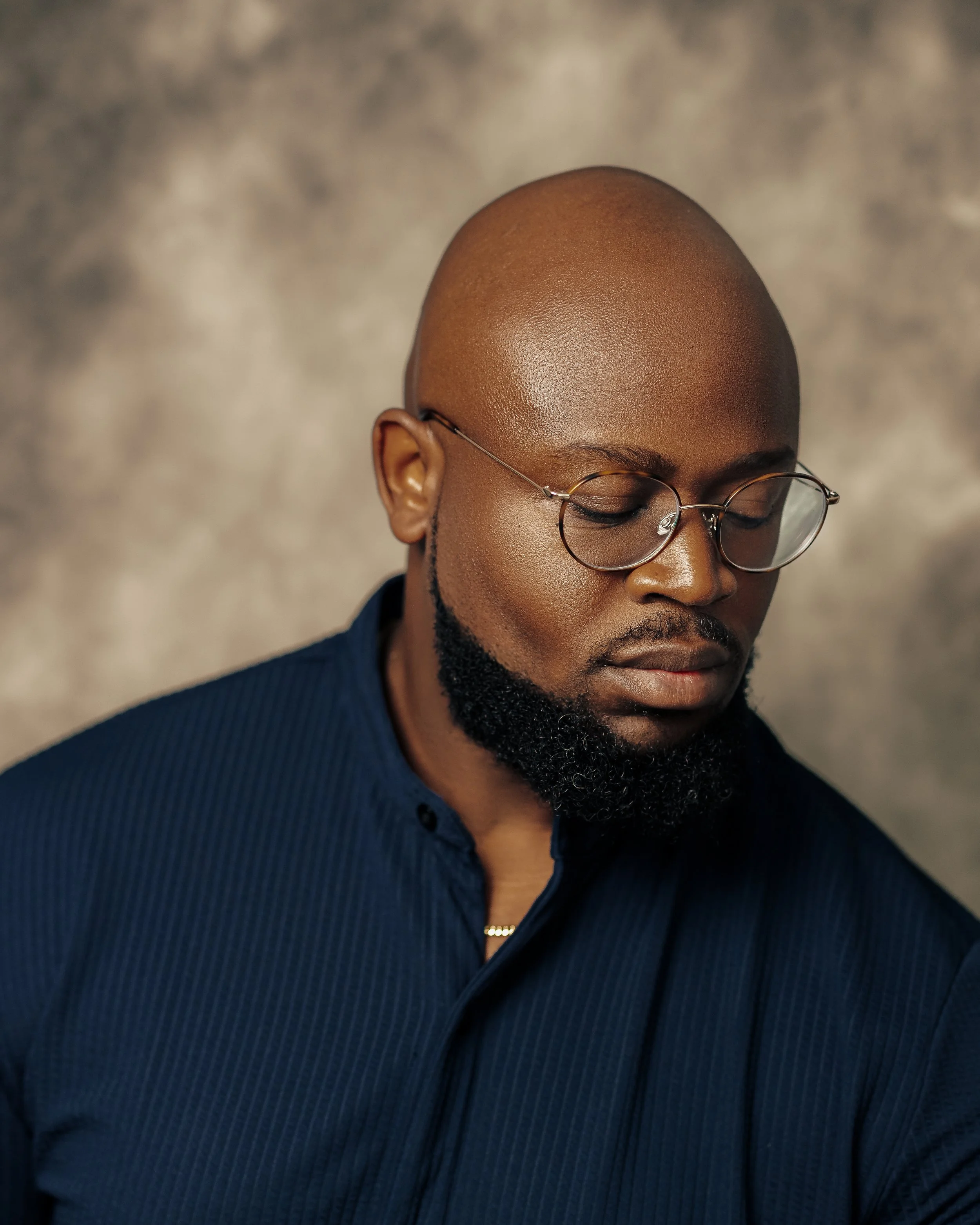 A person with dark skin, a shaved head, glasses with round frames, and a well-groomed beard, wearing a dark, textured blazer, looking downward.