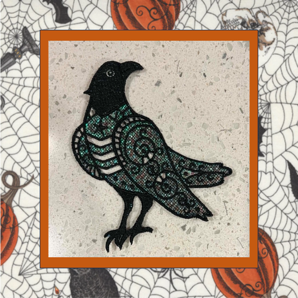 Lace Halloween crow machine embroidery design with shimmering teal Mylar applique and intricate black lace stitching on a light background.
