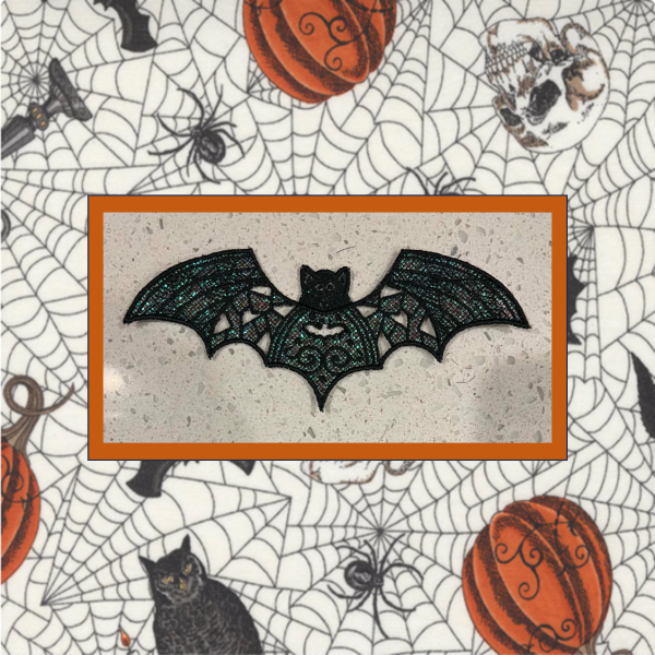 Lace Halloween bat machine embroidery ornament with black lace wings and shimmering green Mylar applique, shown on a spiderweb Halloween fabric background.