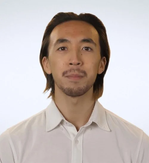 A man with shoulder-length dark hair, a mustache and goatee, wearing a light-colored collared shirt, posing against a plain light background.