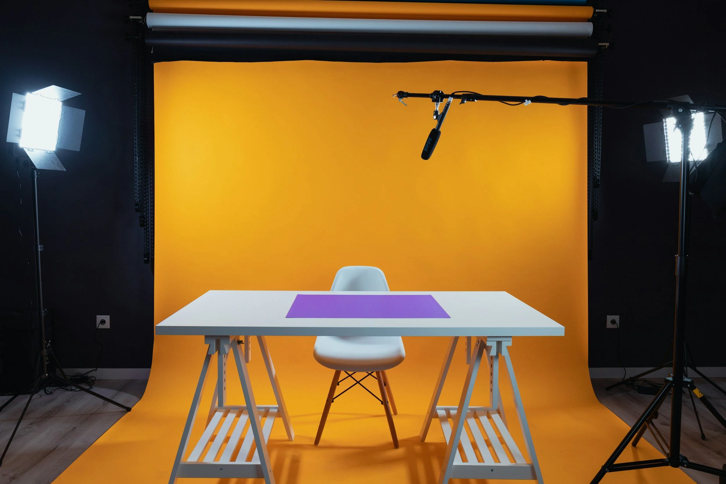 Photography studio setup with a white table, a white chair, a purple rectangular cloth on the table, and yellow backdrop, illuminated by two professional studio lights with softboxes.