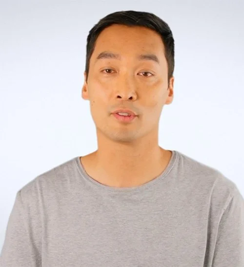 A young man with short black hair wearing a light gray T-shirt, standing against a plain white background.