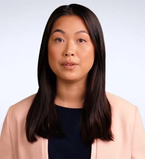 A woman with long dark hair wearing a beige blazer over a dark top, looking directly at the camera against a plain light background.