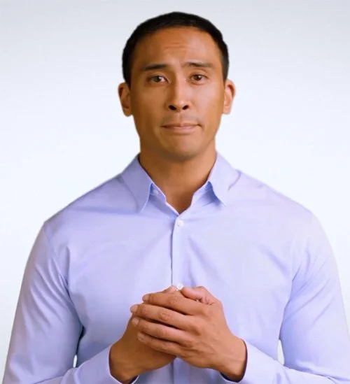 Man wearing a light blue dress shirt, standing against a plain white background, looking directly at the camera with hands clasped in front.