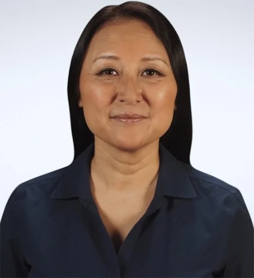 A woman with long black hair wearing a dark blue shirt, smiling slightly against a plain light background.