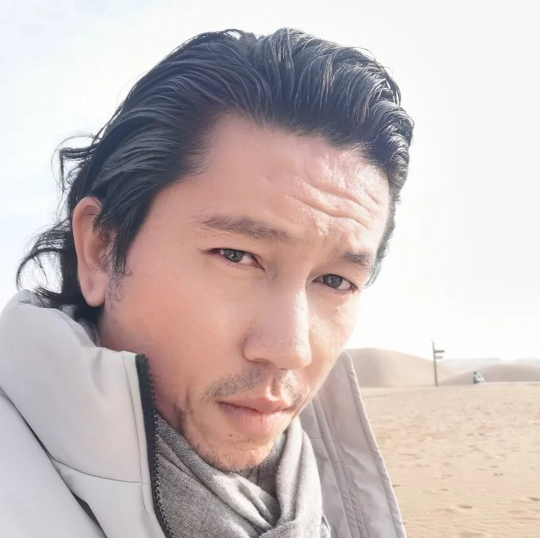 A man with dark, slicked-back hair taking a selfie in a sandy desert with a flag in the background.
