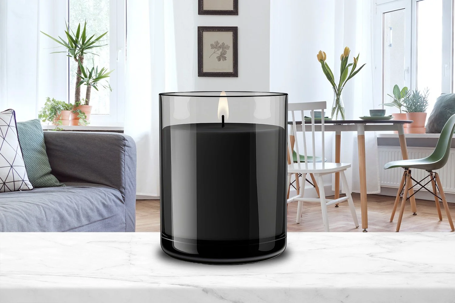 A black candle with a lit flame in a clear glass holder on a white marble surface in a bright living room with a gray sofa, green and patterned pillows, potted plants, and a dining table with chairs.
