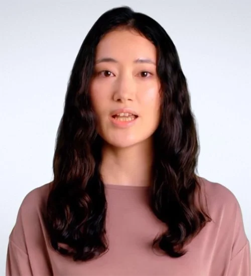A woman with long wavy dark hair and a neutral expression, wearing a mauve top against a plain light background.