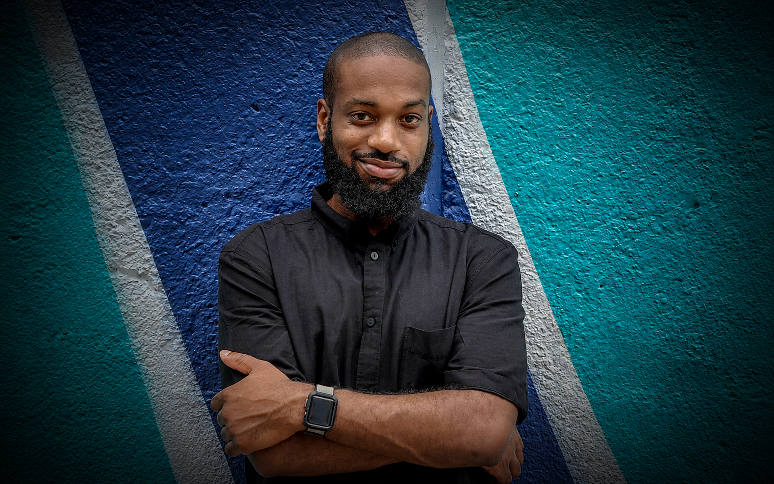A man with a beard and short hair, wearing a black button-up shirt and smartwatch, stands with arms crossed in front of a painted textured wall with blue and teal colors.