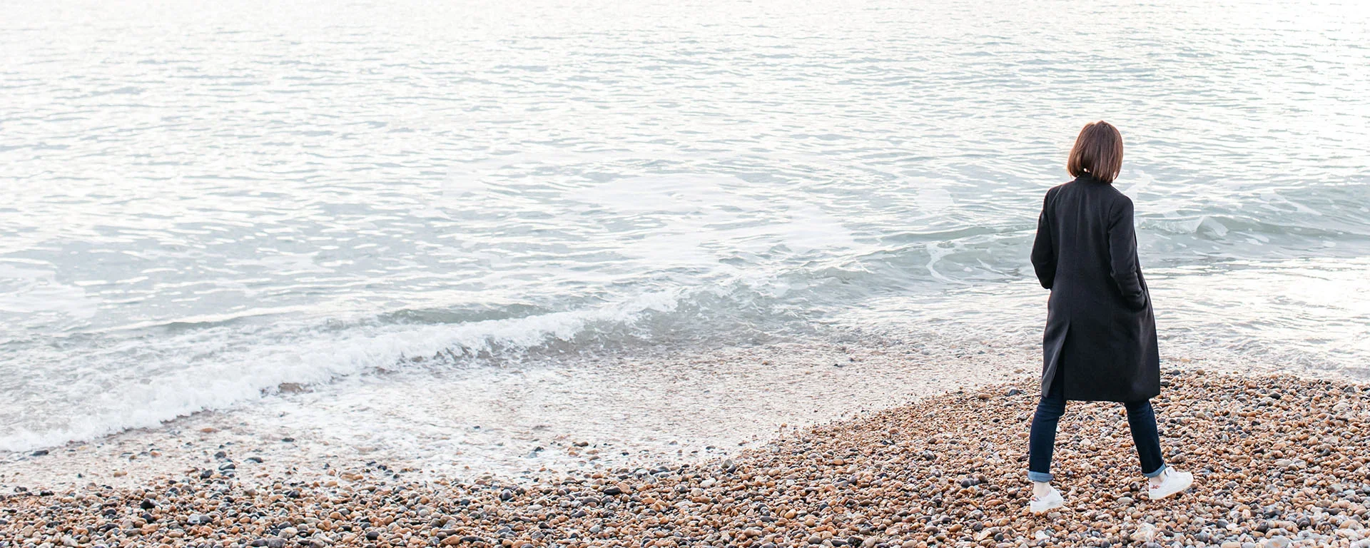 A woman in a black coat and white shoes stands on a pebble beach facing the ocean.