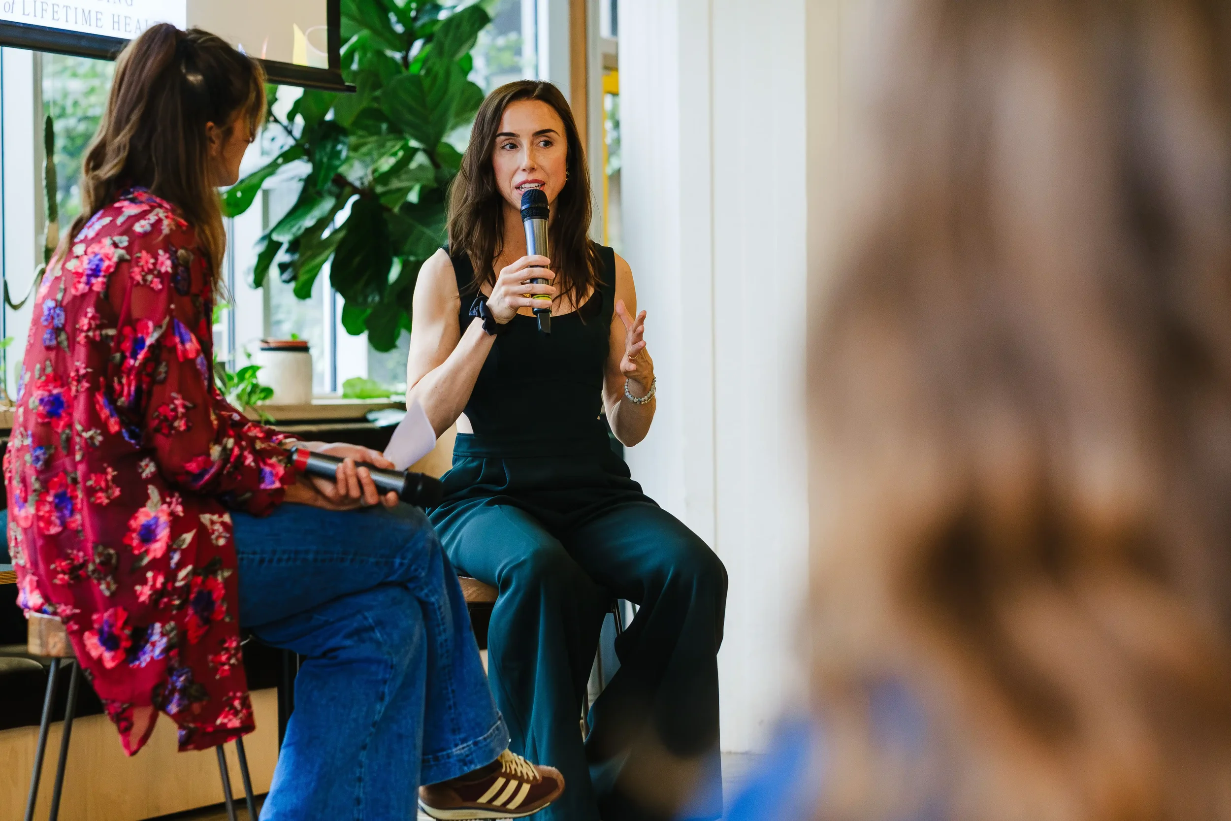 Dr. Jenna in a black sleeveless top and black pants is sitting and speaking into a microphone. Another woman, wearing a red floral top and jeans, holds a microphone and likely moderates the discussion. Green plants and windows are in the background.