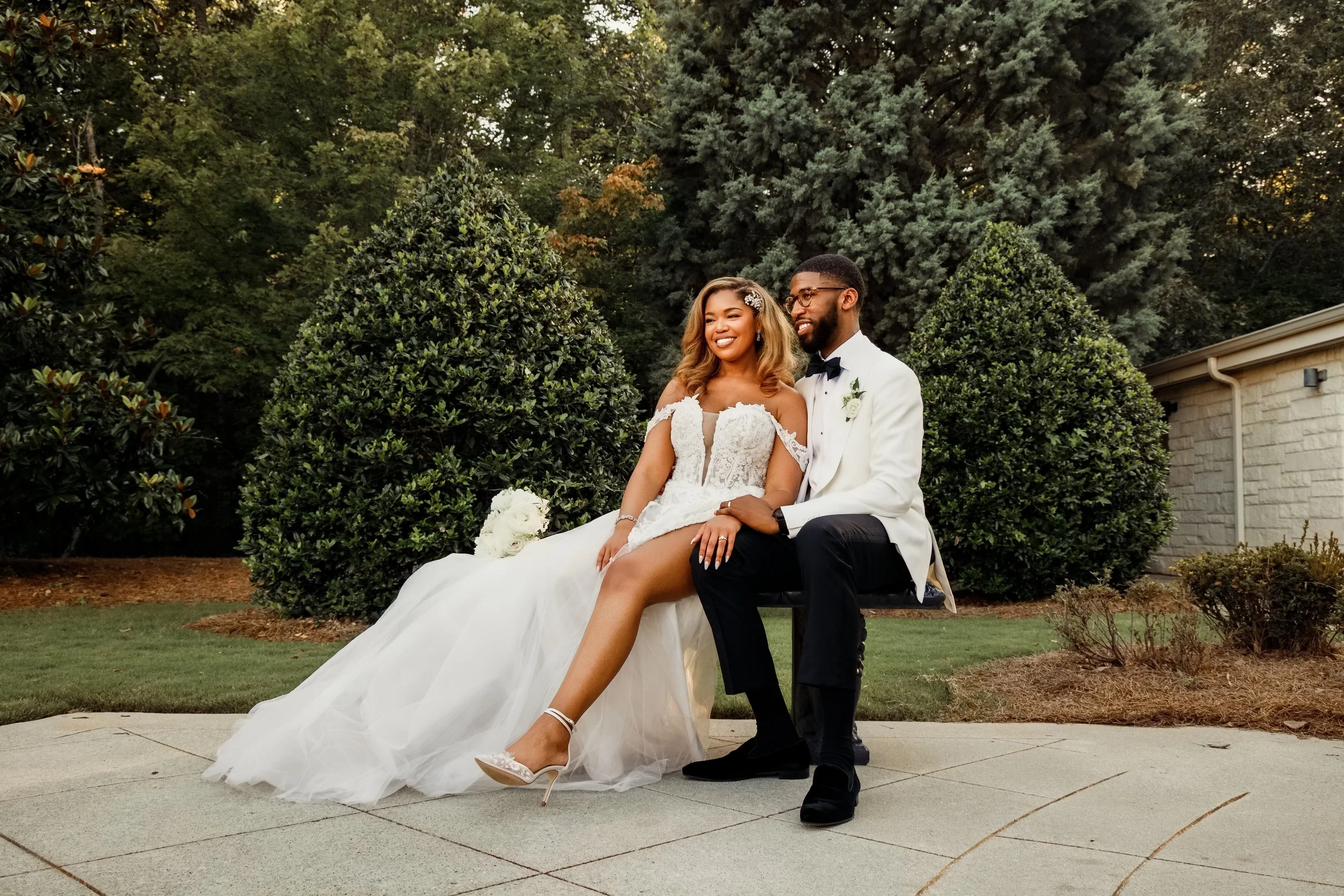 A newlywed couple sitting on a bench outdoors, smiling, surrounded by green trees and bushes, with the bride holding a bouquet of white roses.