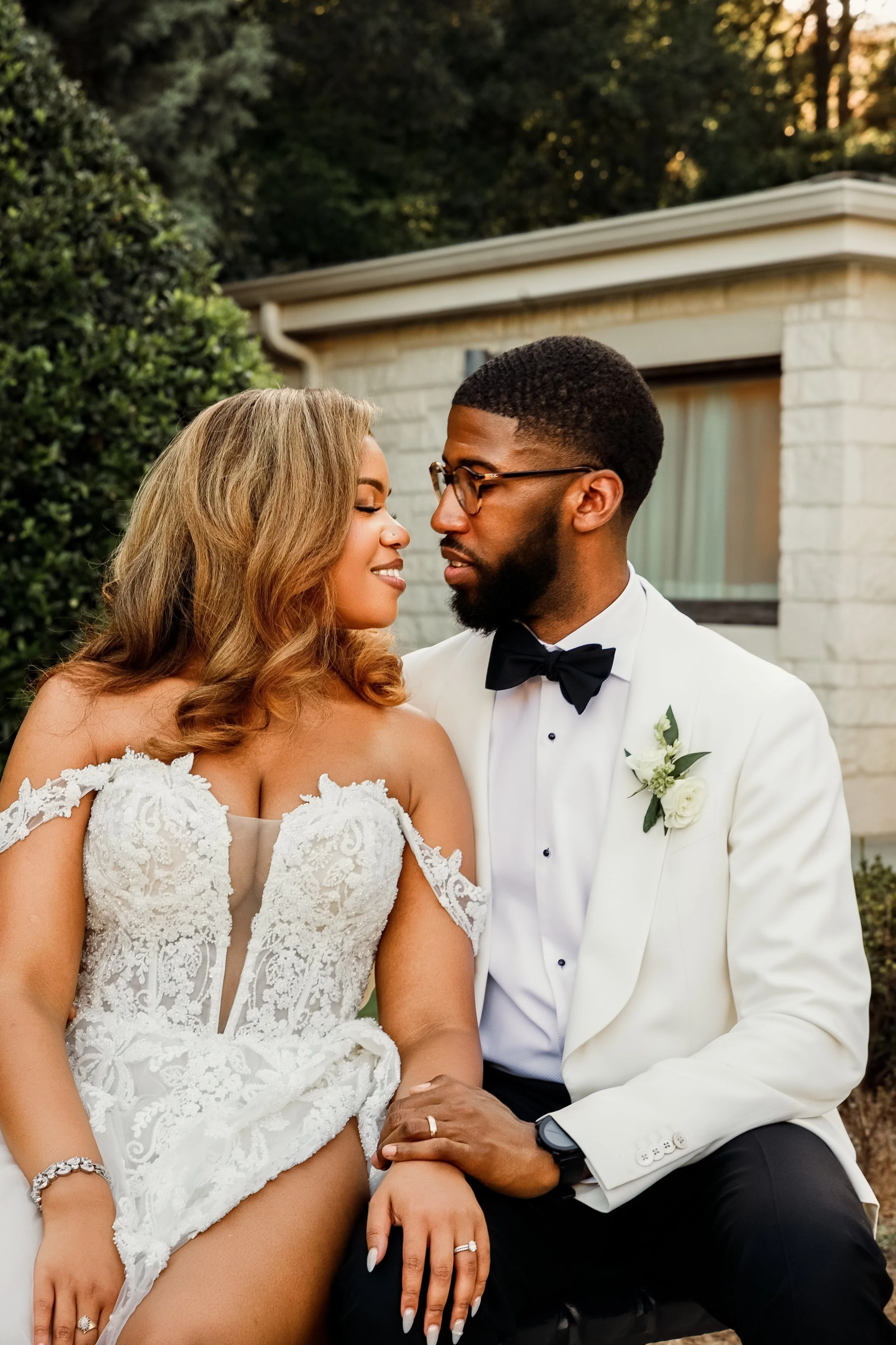 A bride and groom sitting closely together outdoors at sunset. The bride wears a white lace wedding dress and has long wavy hair. The groom wears a white tuxedo with a black bow tie, glasses, and a boutonniere. They are holding hands and looking into