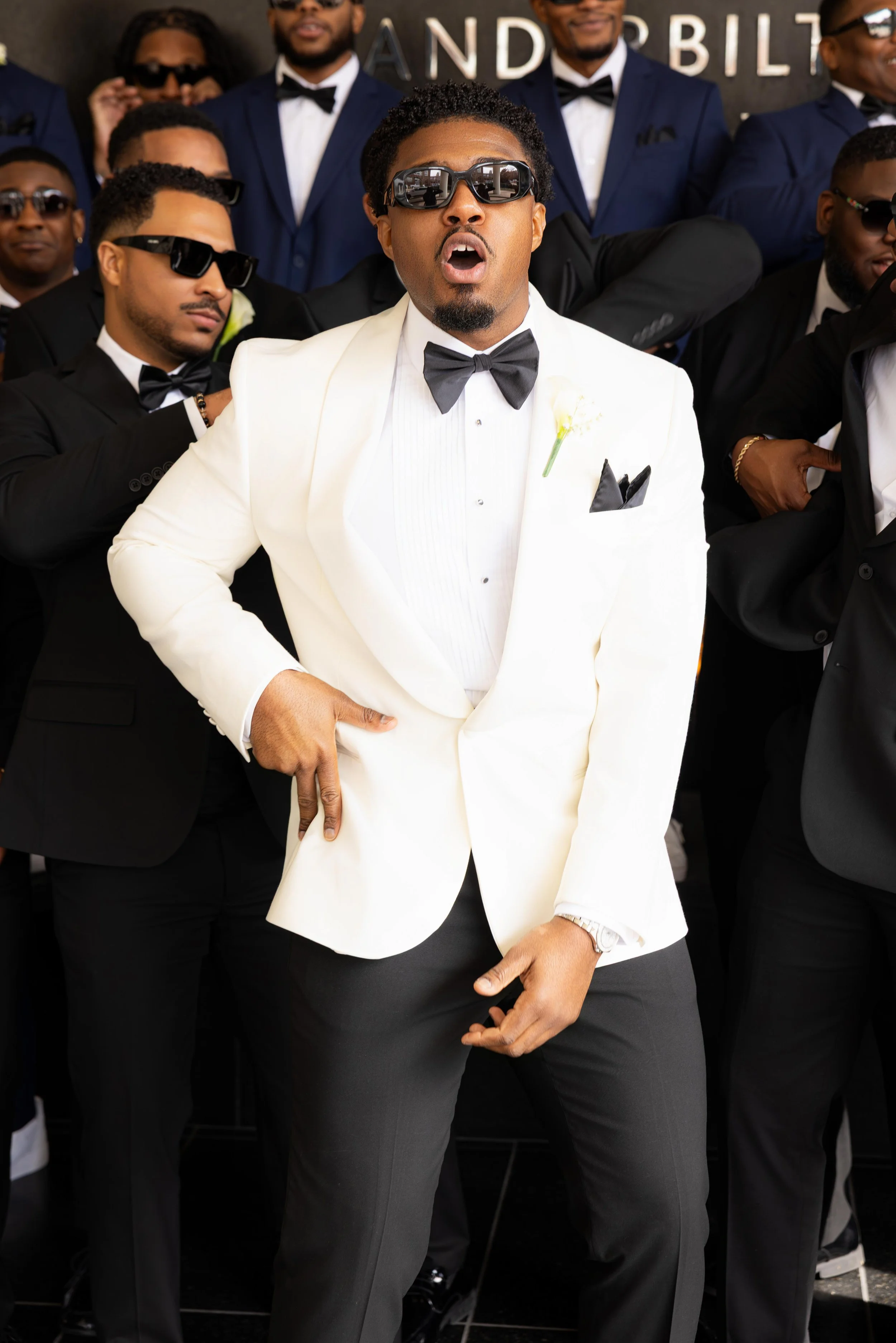 A group of men dressed in tuxedos and sunglasses, posing at a formal event. The man in front is wearing a white tuxedo and appears surprised or expressive.