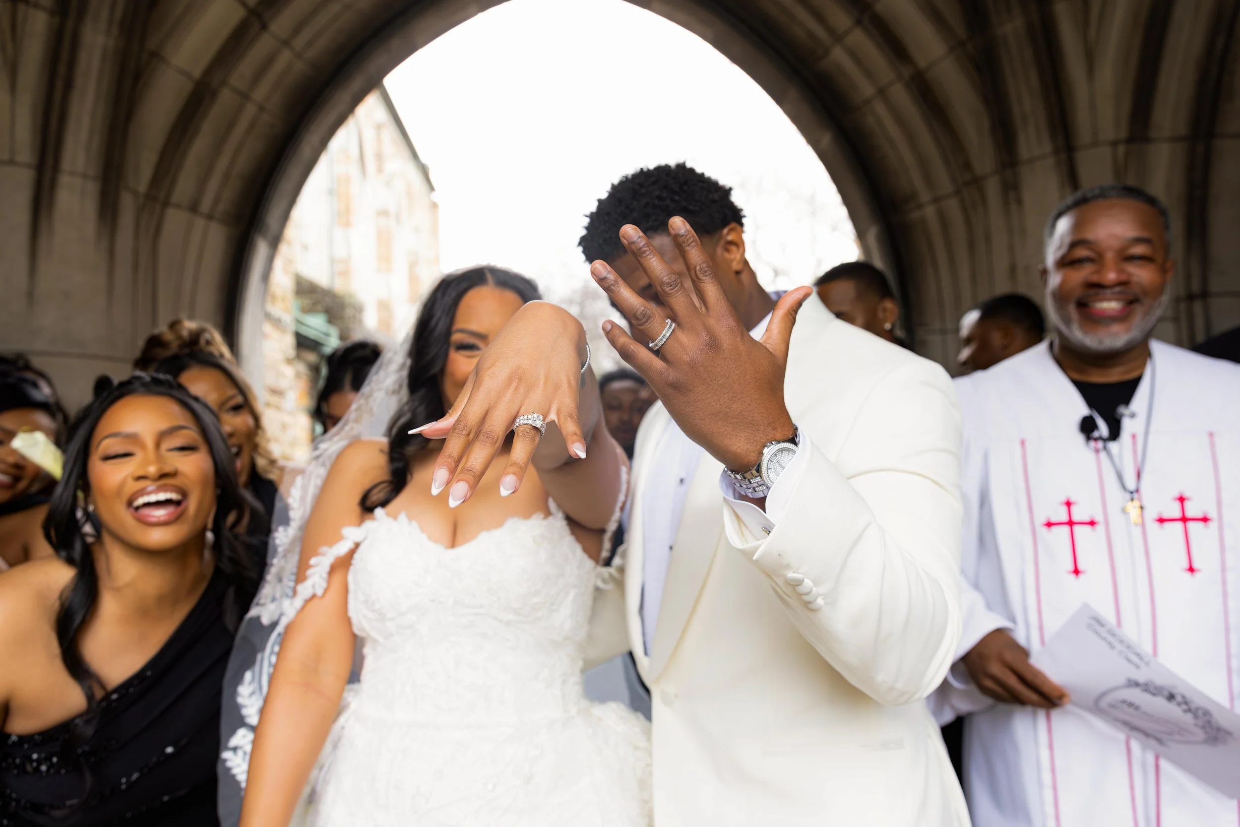 A bride and groom display their wedding rings during their wedding ceremony, surrounded by joyful friends and family in an outdoor setting under an archway.