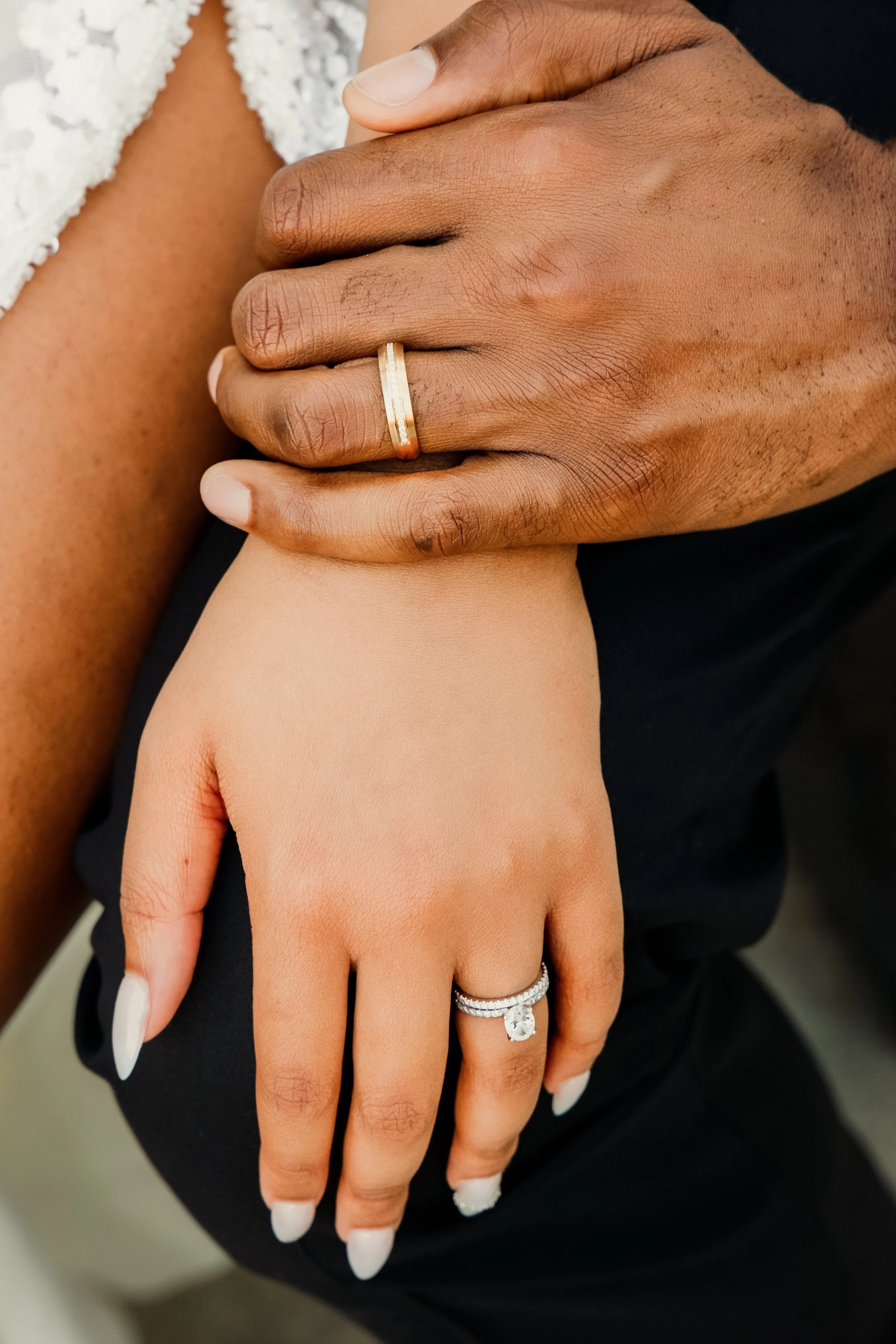 Close-up of a person’s hand with a wedding ring, resting on another person’s hand, with another ring visible on the other person’s finger. The background shows part of a white lace garment.