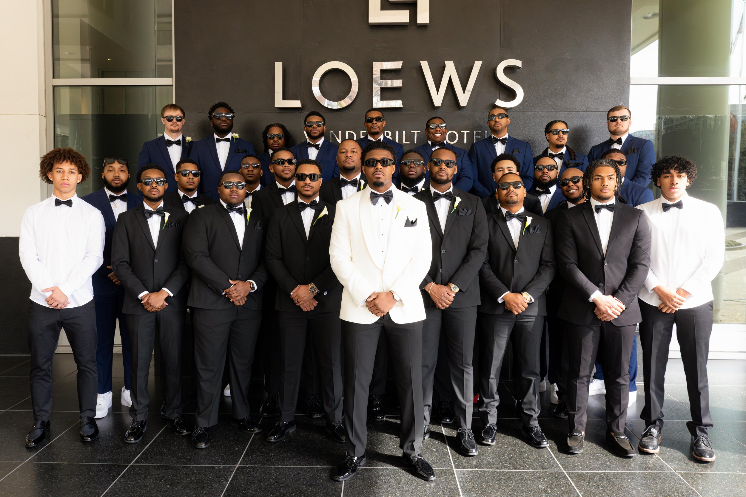 A group of men dressed in tuxedos and suits with sunglasses, standing in front of a sign that reads 'LOEWS' at the underlit hotel. Some are smiling, and some are serious, standing on a black tiled floor.