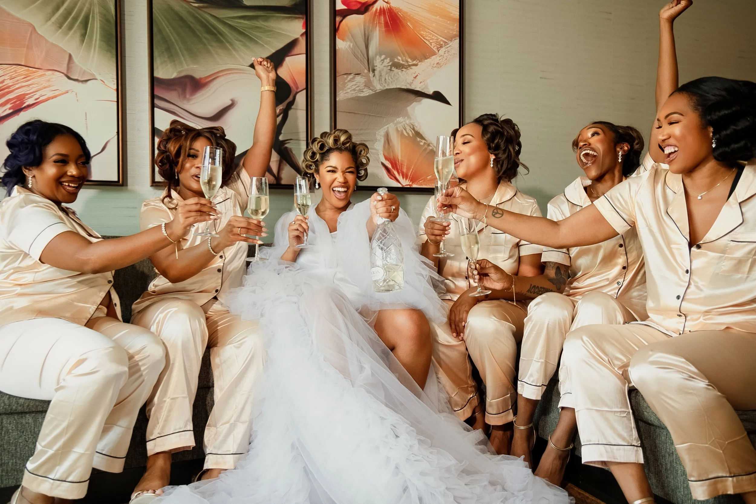 Six women in matching beige pajamas celebrating with champagne in a living room, one woman dressed as a bride with curlers in her hair and a white gown, and they are smiling and laughing.