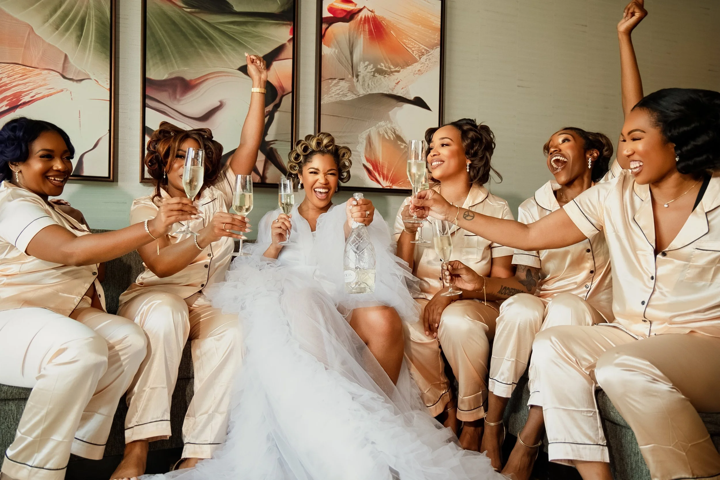 A group of women, dressed in matching satin pajamas, celebrating around a woman in a wedding dress, who is holding a bottle of champagne, with others holding glasses of champagne and cheering in a decorated indoor setting.