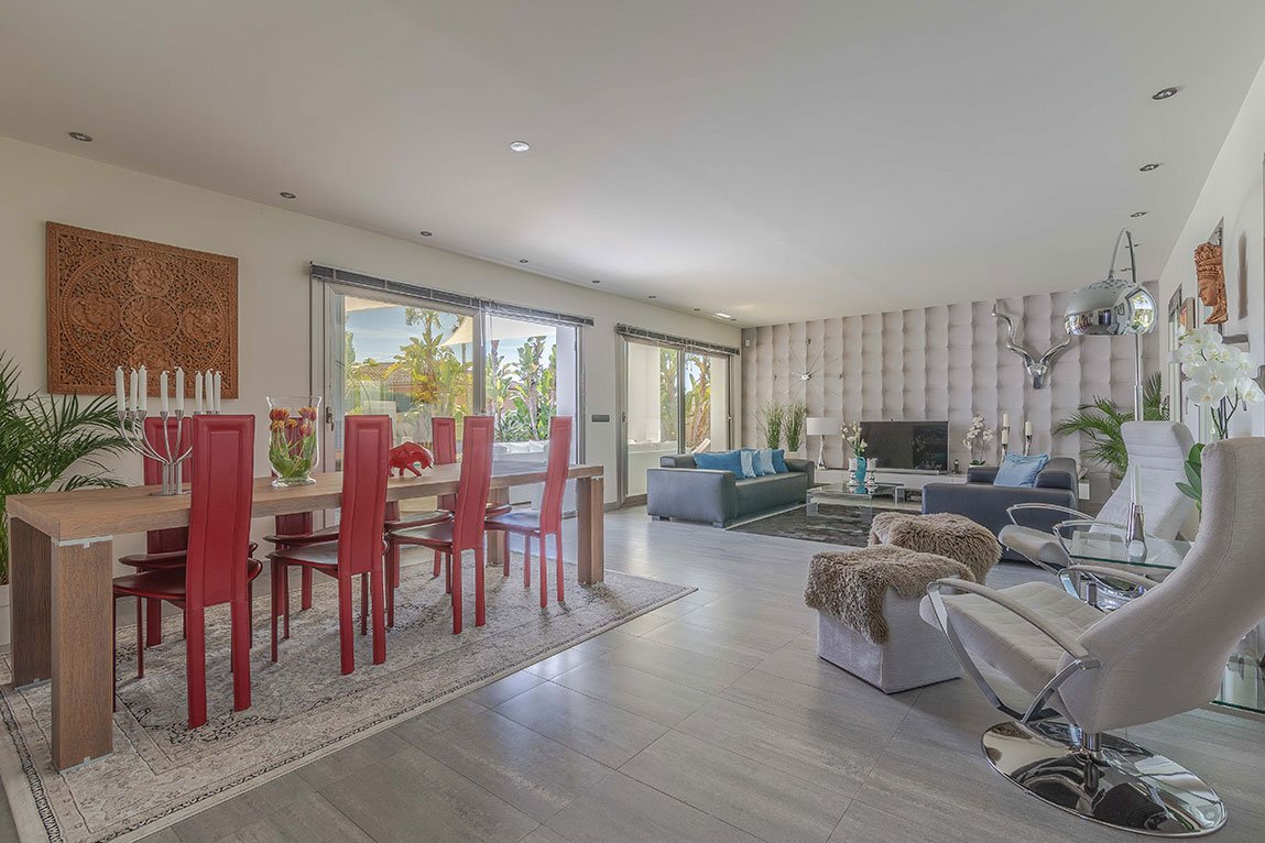 Sala de estar con muebles modernos, sillones grises y blancos, televisión, paredes acolchonadas, decoraciones florales, y ventanas que muestran árboles y plantas