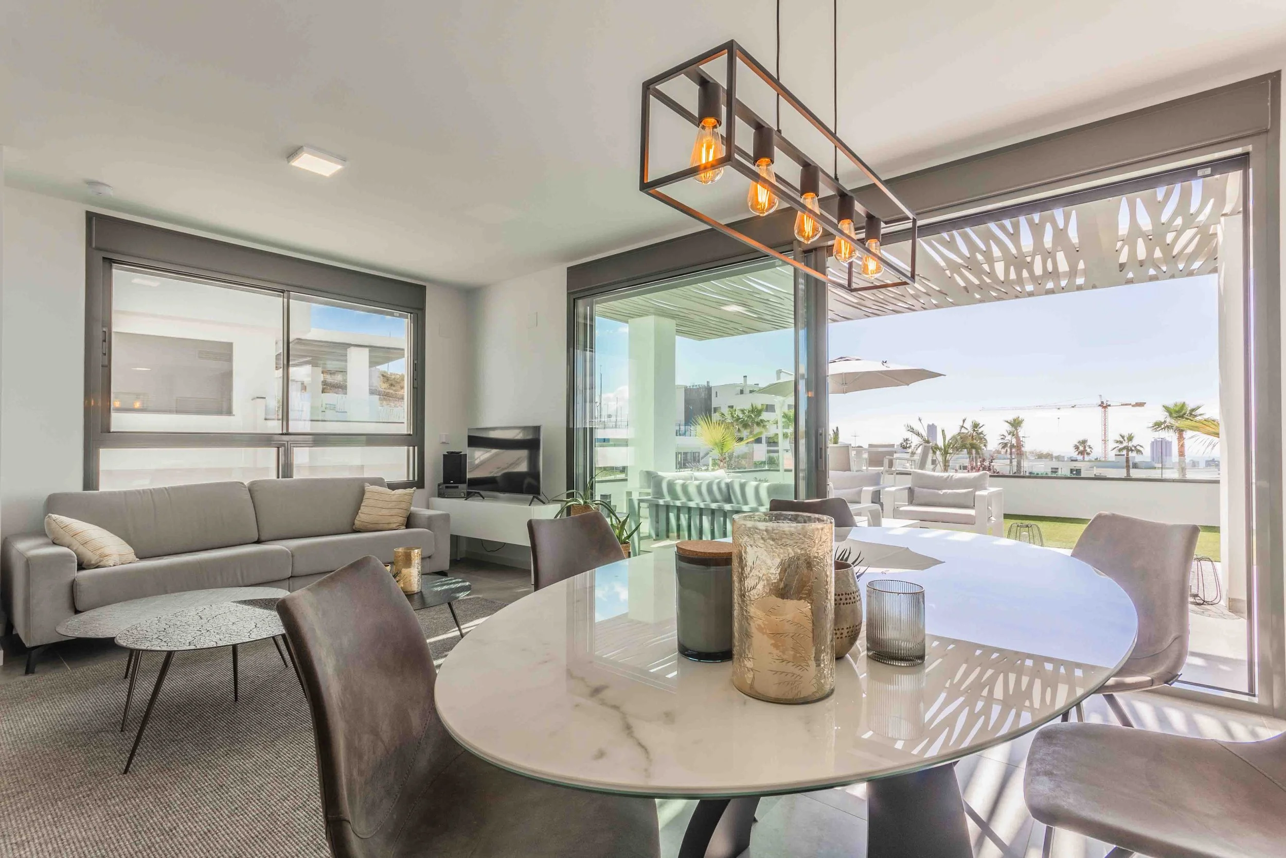 Sala moderna con sillones grises, mesa de comedor ovalada de mármol, decoraciones en tonos neutros, ventanas grandes y vista a un balcón con sombrilla y palmeras.