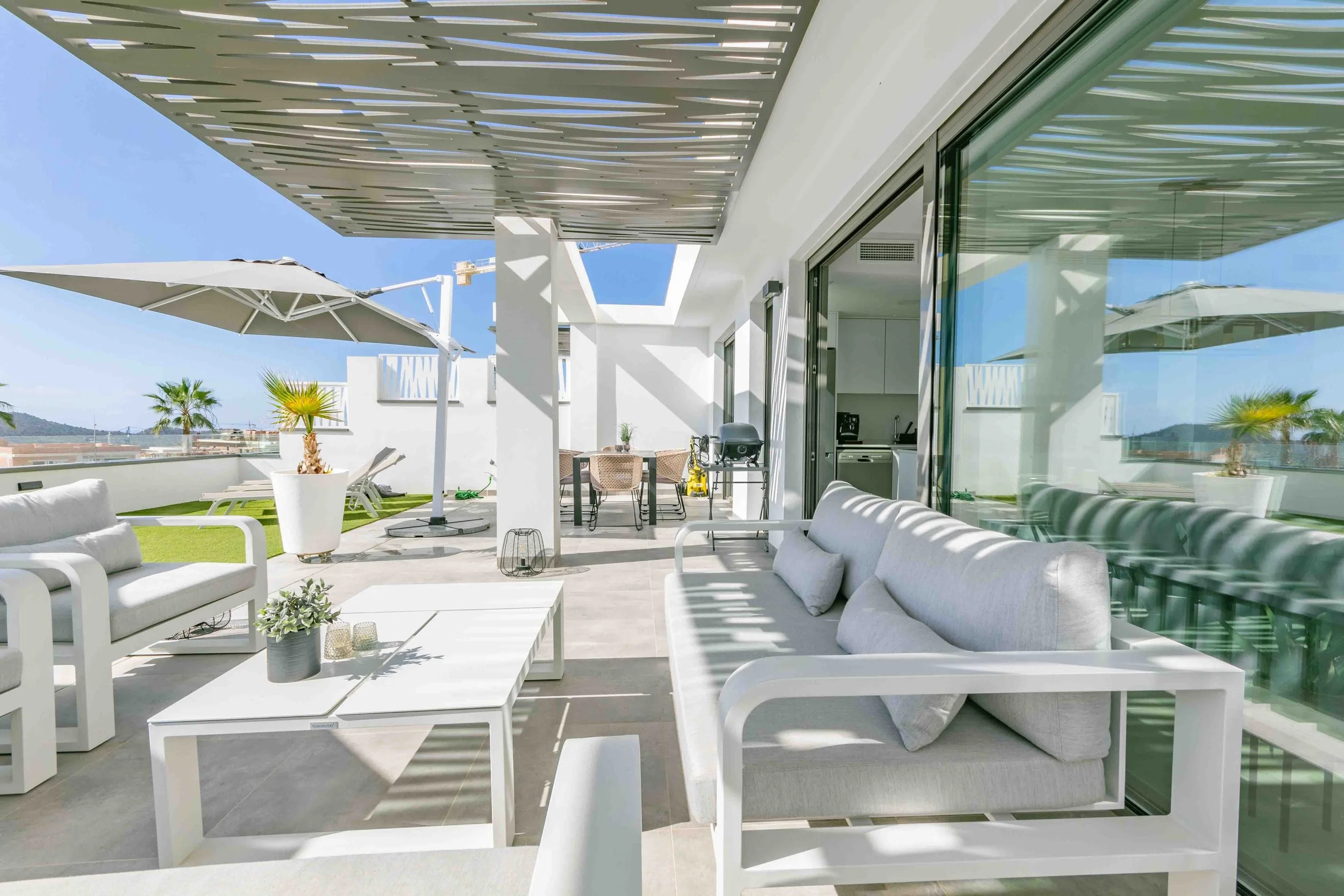 Terraza moderna con muebles blancos, sombrillas, plantas y vista al cielo y montañas al fondo.