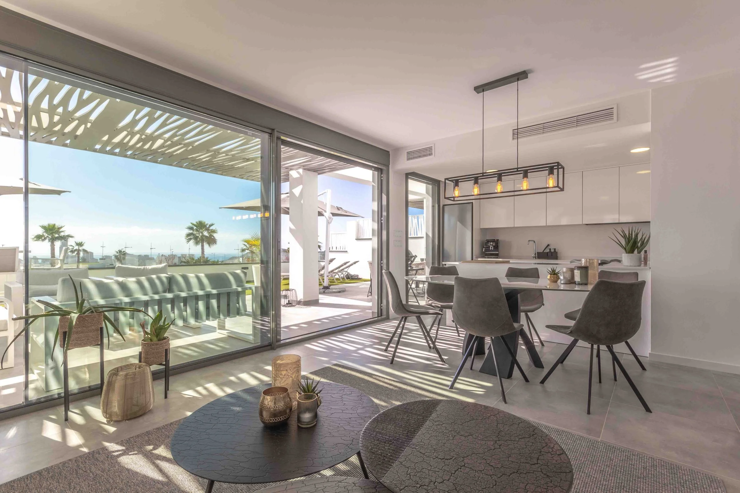 Sala contemporánea con comedor y cocina, grandes ventanales con vista al exterior, plantas decorativas, mesa y sillas modernas, lámpara colgante, terraza exterior con sombrillas y palmeras.