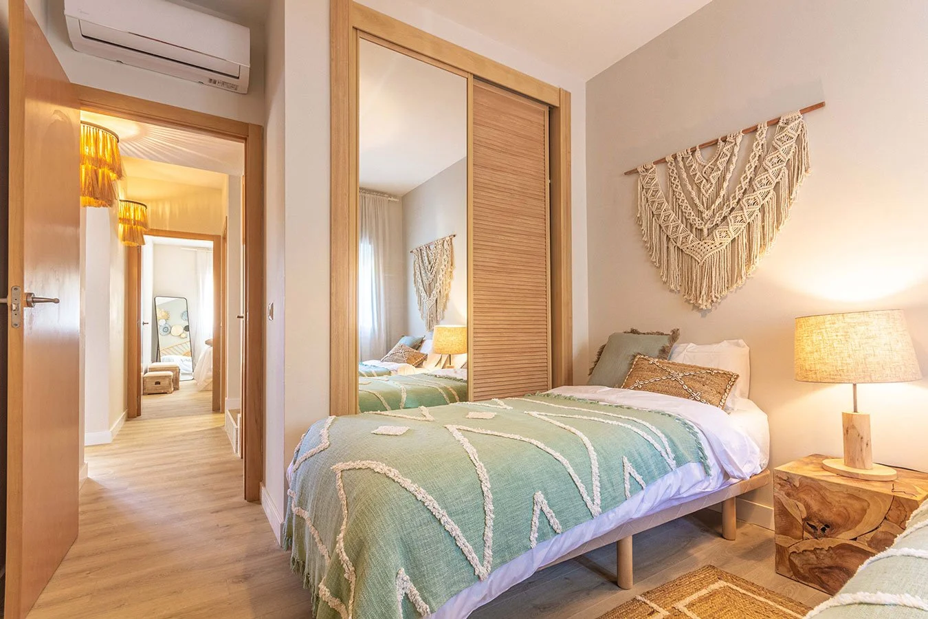 Dormitorio con dos camas, decorado en tonos neutros y verdes, paredes blancas, papel tapiz con macramé en la pared, lámparas de mesa de madera, closet con puertas corredizas de madera, y detalles decorativos como cojines y alfombra en tonos tierra.