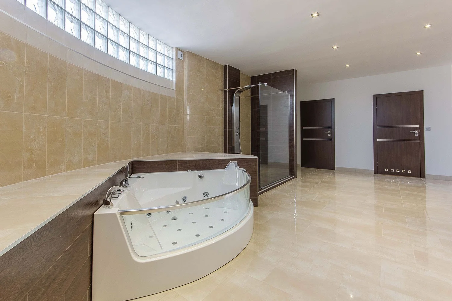 Baño amplio con tina de hidromasaje, ducha con mampara de cristal, paredes de azulejo beige, suelo de baldosas beige, puertas de madera oscura, ventanas de bloques de vidrio, luces empotradas en el techo.