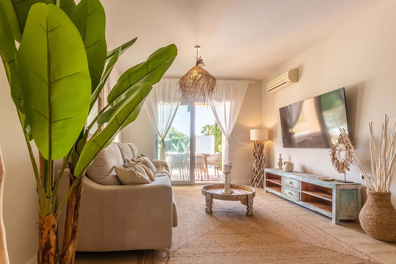 Sala de estar con sofá beige, mesa de centro de madera clara, televisor montado en la pared, lámpara, decoraciones de estilo rústico, plantas y puerta con vista a un balcón con vista al exterior