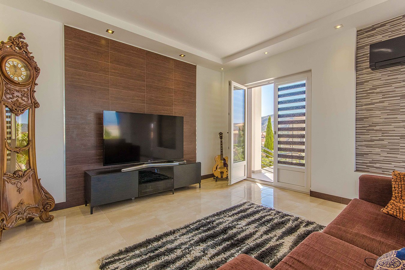 Sala de estar con pared de madera, televisor, reloj antiguo de pared, guitarra junto a la ventana, sofá marrón y alfombra de felpa