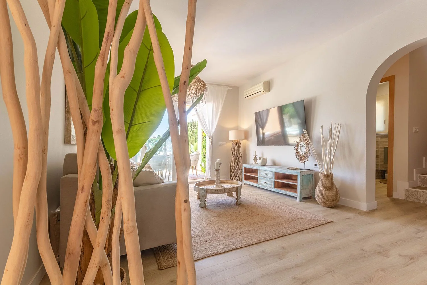 Salón decorado con muebles de estilo rústico y plantas grandes, con paredes blancas, suelo de madera clara y una televisión montada en la pared.