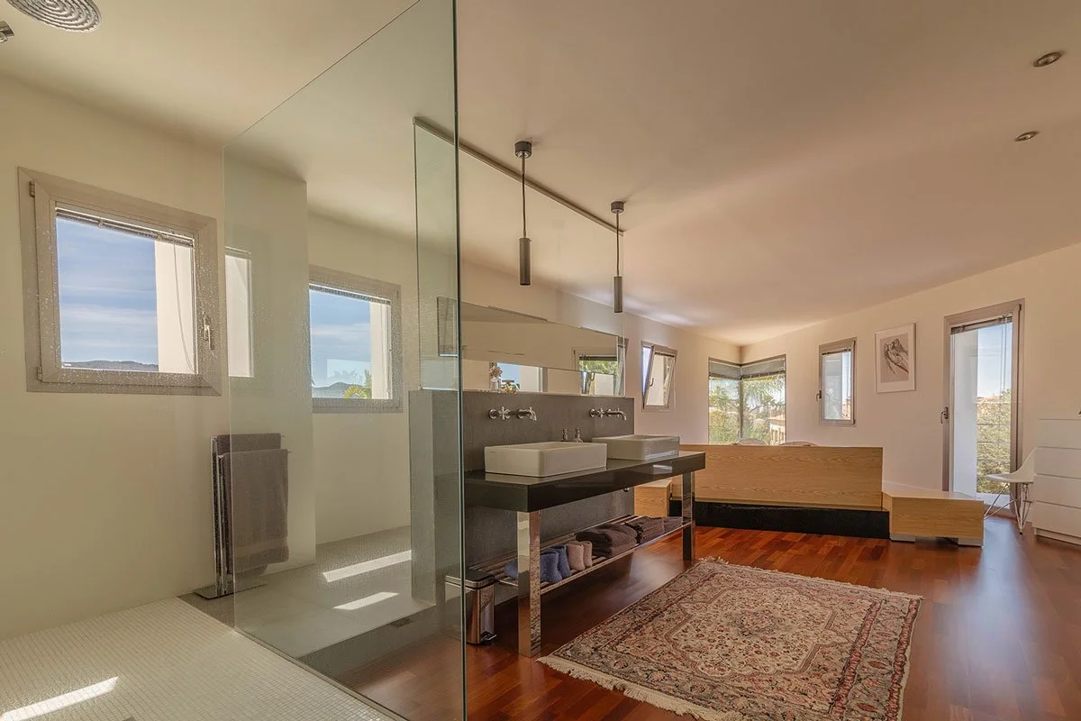 Habitación moderna con balcón, paredes blancas, ventanas grandes, suelo de madera, y un área de lavabo con dos lavabos y espejo.