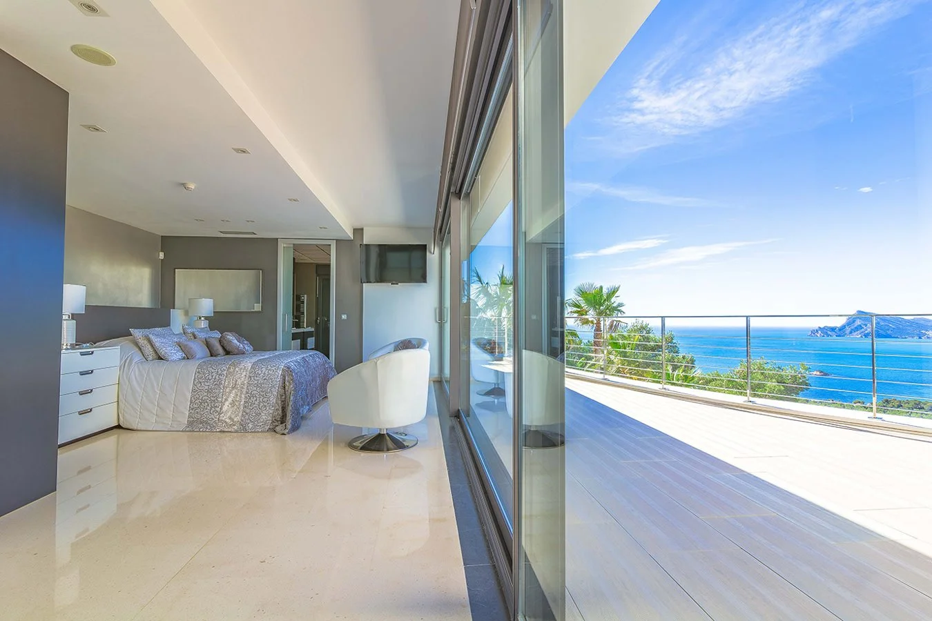 Habitación moderna con cama, sillas blancas y vista al mar desde un balcón amplio