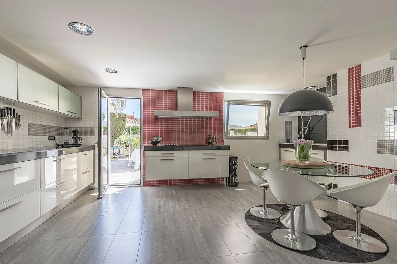 Cocina moderna con pared de azulejos rojos y blancas, mesa de comedor ovalada con sillas blancas y luz colgante negra, puertas y ventanas abiertas hacia un jardín exterior.