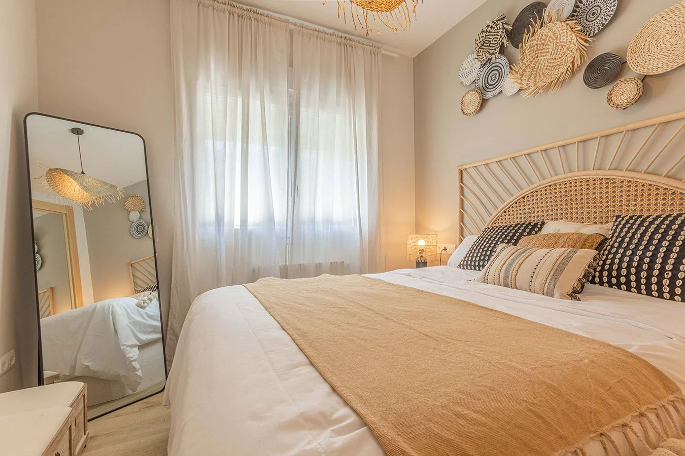 Habitación decorada con cortinas blancas, pared con arte en cestería, cama con cabecera de madera y almohadones en tonos negró y beige, lámpara de mesa y espejo de pie