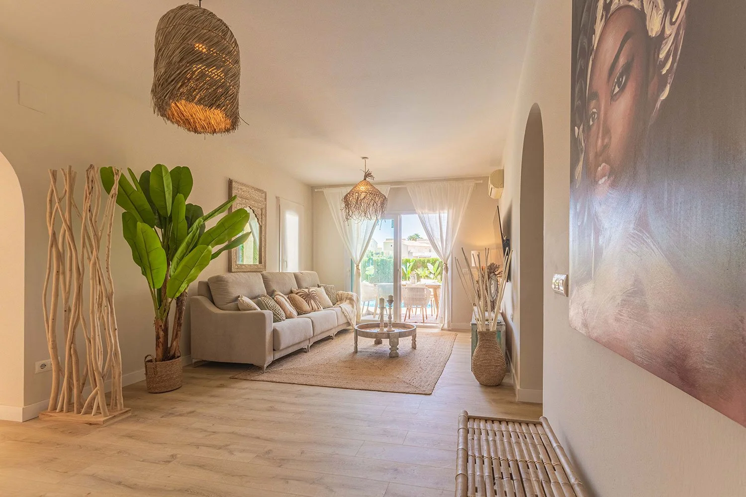 Salón de estilo tropical con sofá beige, plantas grandes, cortinas blancas, alfombra, mesa de centro y decoración natural.