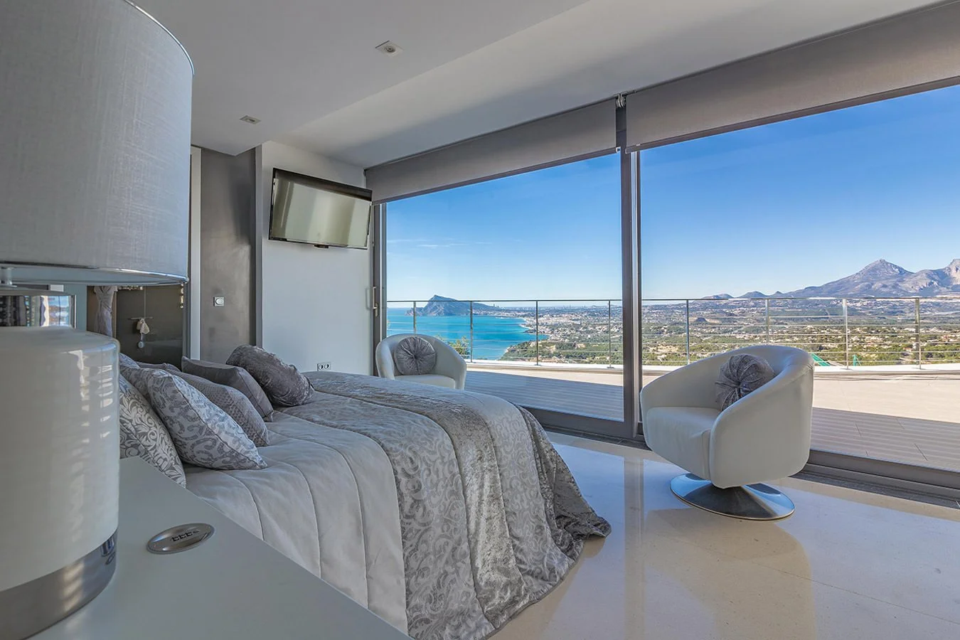 Habitación moderna con vista panorámica del mar y montañas, cama con ropa de cama en tonos plateados y dos sillas blancas con cojines decorativos.